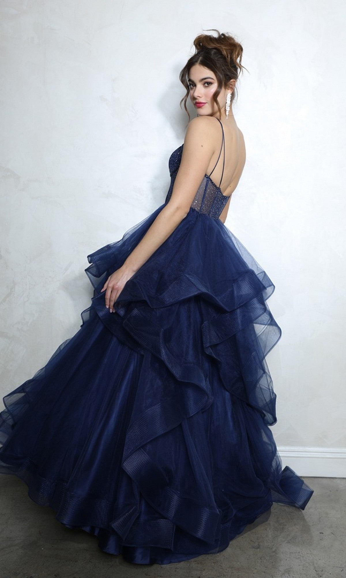 A woman wearing the Eureka 9950 long prom dress, a dark blue layered tulle ballgown, stands against a light wall, looking over her shoulder with her hair styled up.
